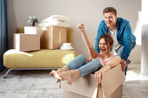 Young family couple bought or rented their first small apartment. Cheerful woman scream sitting in box. Guy move her. They play game during moving in. Local movers